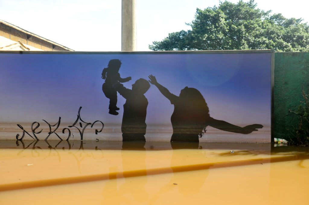 Imagem da silhueta de um casal com seu filho pequeno pintada em um muro que está sob a enchente do Rio Grande do Sul. Jornal UFRGS - Crédito Flavio Dutra