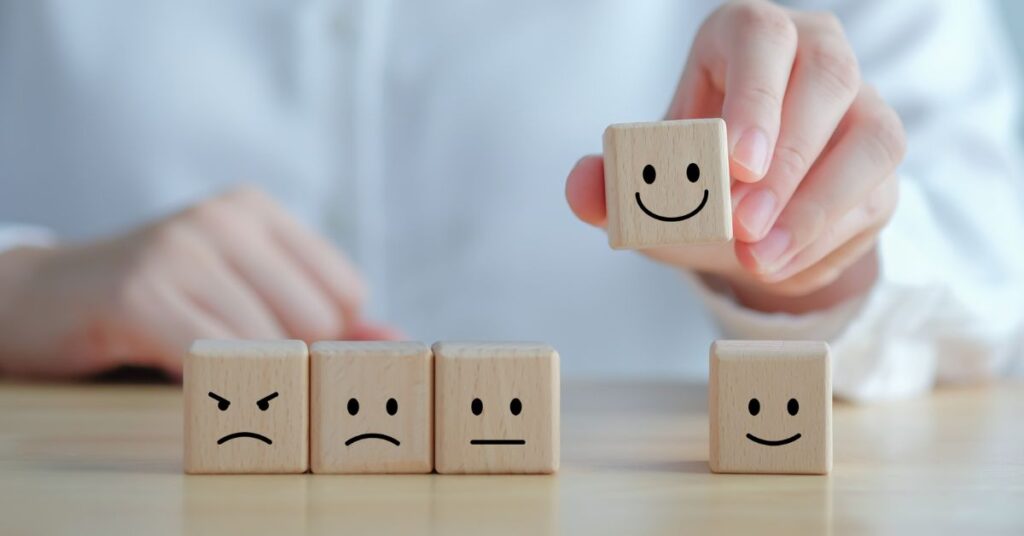 Mãos de uma pessoa branca inserindo um quadradinho de madeira com um sorriso desenhado entre outros quatro quadradinhos com, três com carinha triste e um outro sorrindo.