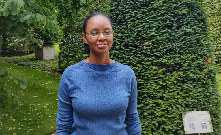 Física Rita de Cássia,mulher negra, usando blusa azul, de lã e mangas longas, em pé em um campo verdejante.