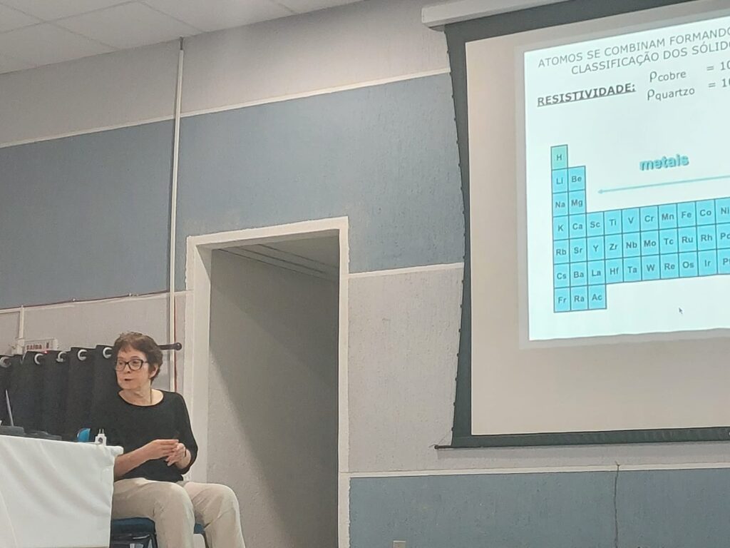 A física, professora Belita Koiller, uma senhora de cabelos loiros escuros, curtos, usando camiseta preta. Ela está sentada e fazendo uma apresentação durante 1⁰ Workshop de Física da Matéria Condensada Teórica do Rio de Janeiro.