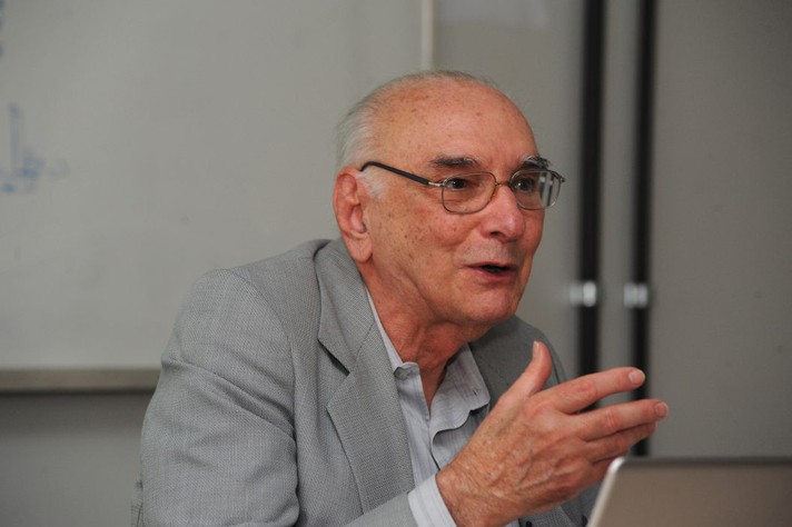 Professor de Física Ramayana Gazzinelli, um senhor branco de 80 anos, vestindo terno cinza e camisa branca, usando óculos, falando e gesticulando, sentado, durante uma entrevista.