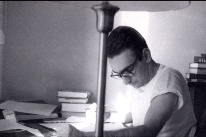 Foto da década de 1960 do jovem pesquisador, Ramayana Gazzinelli estudando, sentado atrás de uma escrivaninha em uma sala cheia de livros,