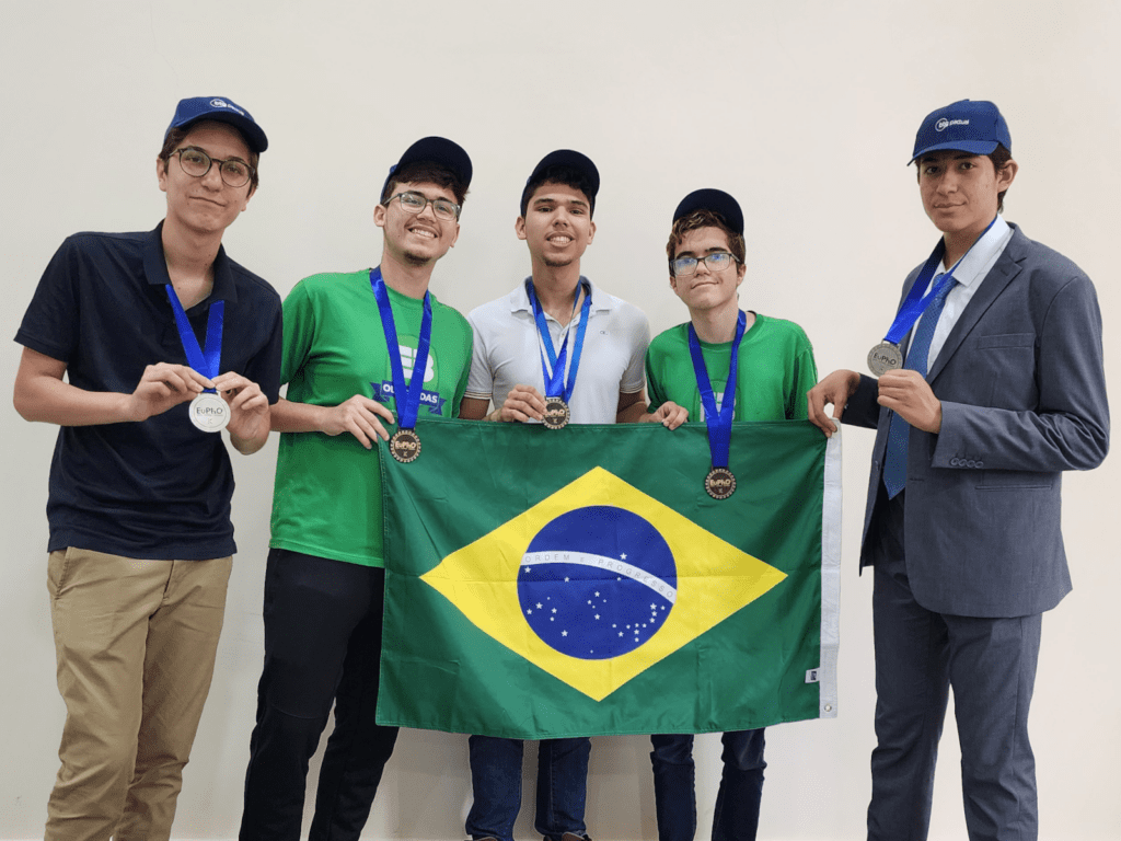 Quatro alunos do ensino médio e um professor orientador, posando com a bandeira do Brasil durante a EuPhO 2024.